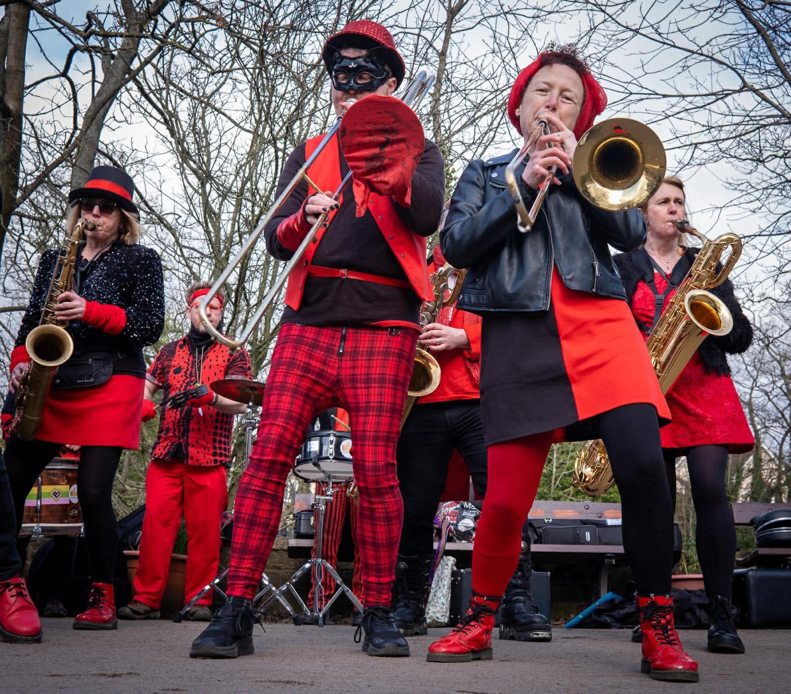 A vibrant band performing outdoors, featuring musicians dressed in red and black attire. They are playing various brass instruments, including trumpets and trombones, with a lively atmosphere created by their energetic expressions and coordinated outfits.