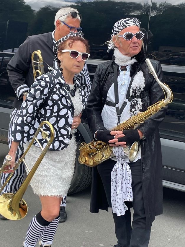 A group of musicians in quirky black and white outfits, including a woman with a trombone and a man holding a saxophone, standing next to a black vehicle.