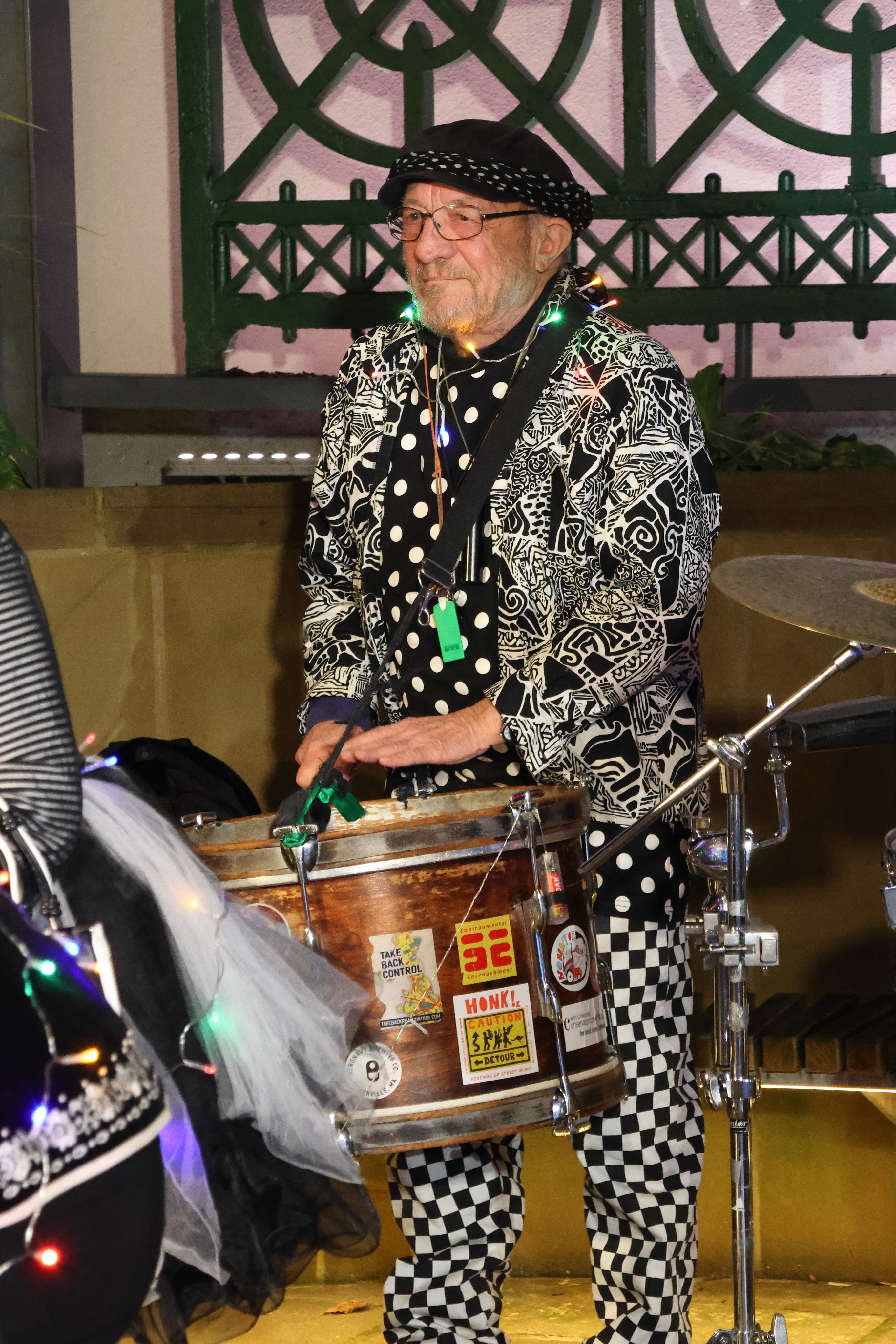 An elderly musician wearing a patterned black and white outfit and a cap plays a drum, adorned with colorful lights.