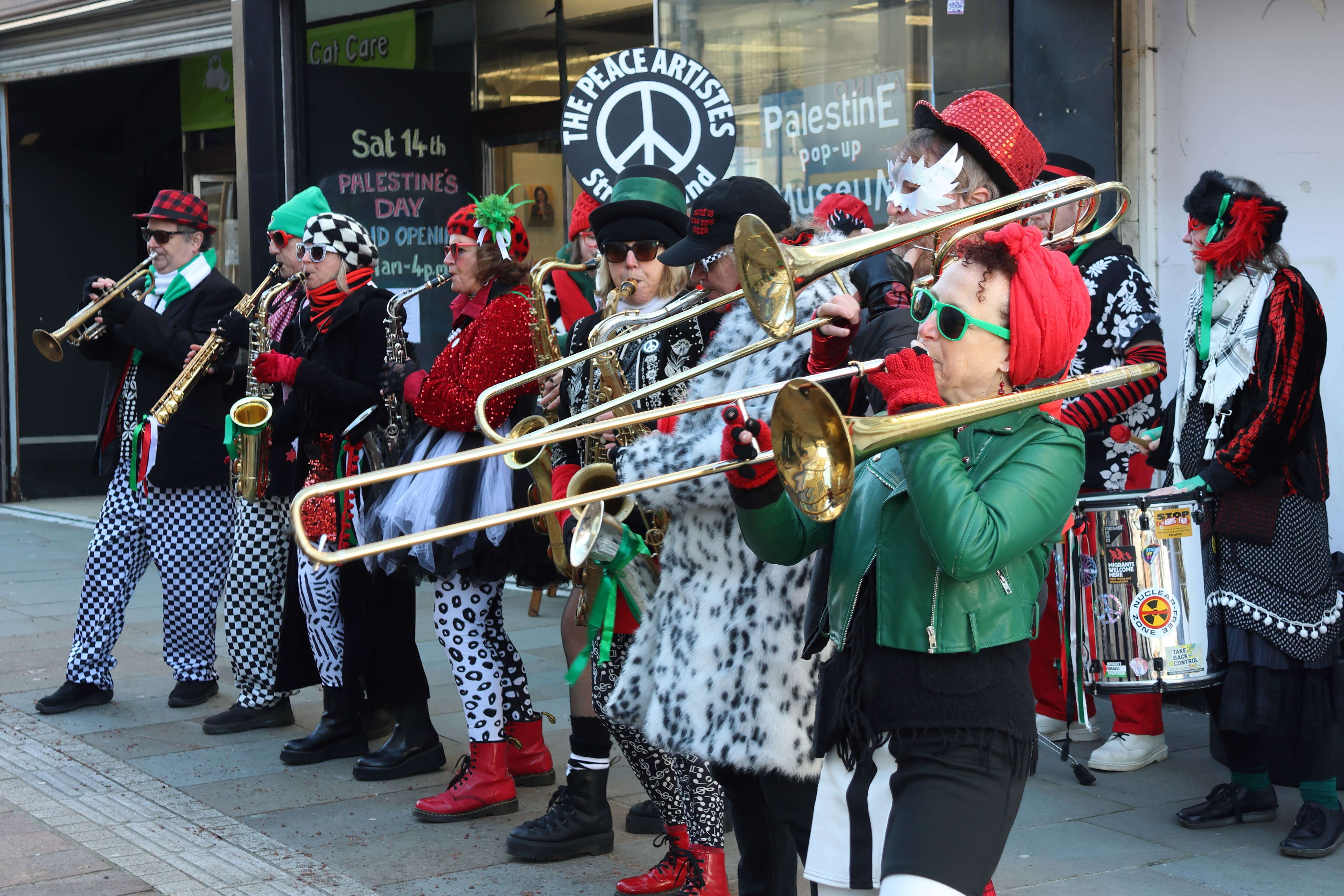 A lively group of musicians wearing colorful costumes and accessories, playing brass instruments on a street. The musicians are adorned with sequins, hats, and patterned clothing, creating a festive atmosphere.