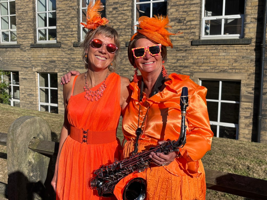 Two women dressed in bright orange outfits, one holding a saxophone, with a building in the background.