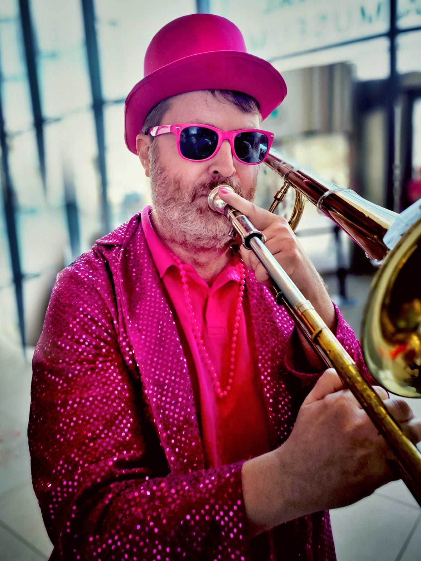 A man wearing a bright pink top hat, sunglasses, and a sequined jacket plays a trombone. He has a beard and is dressed in various shades of pink, standing indoors with a modern architectural background.