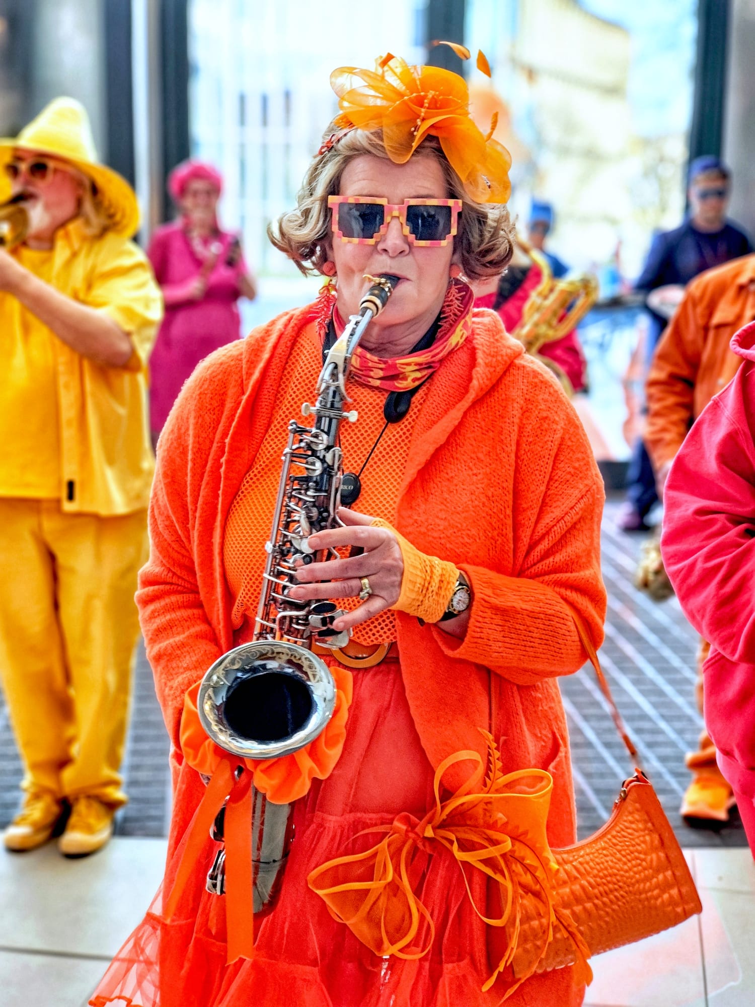 A woman in vibrant orange attire plays the saxophone, wearing oversized sunglasses and a decorative orange hat with a feather. She is surrounded by musicians dressed in bright colors, creating a lively atmosphere.