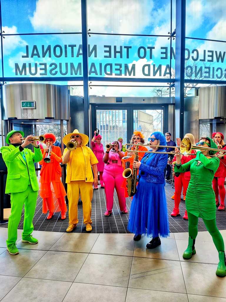 A colorful group of musicians performing inside a modern building, wearing bright outfits in various colors, playing trumpets and saxophones. The large entrance features a sign welcoming visitors to the National Media Museum, with blue skies visible through the glass.